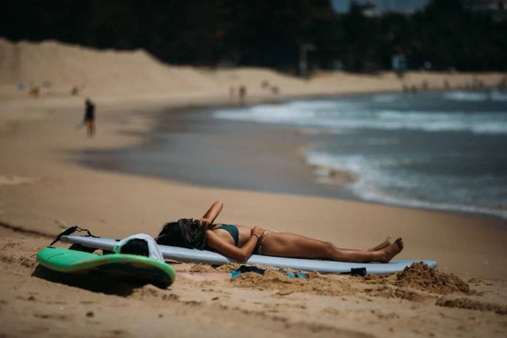 女孩在沙滩上晒太阳，旁边有人把身子埋进了沙子里，只露出一颗头<br>