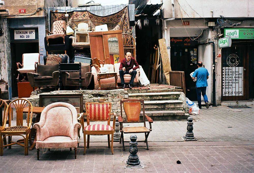 Goksun街头的二手店外，家具像小山一样被堆积在路边。店主坐在沙发上，阴郁沉默。<br>
